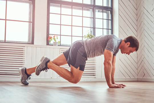 Handsome Muscular Man In A T-shirt And Shorts Doing Functional Exercises On Floor At Home.