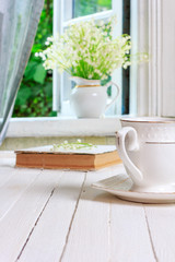 a Cup of tea or coffee and a book on a white wooden vintage retro table and a bouquet of Lily of the valley flowers on the windowsill by the open window in the rustic house in the spring morning