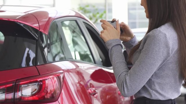 Cropped shot of a happy long haired woman smiling cheerfully using her smart phone taking photos of her new automobile. Female customer buying a car at the dealership showroom.
