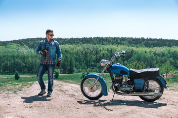Portrait young guy with sunglasses and Jeans jacket uses a smartphone, holds in hands, vintage retro motorbike. biker wearing jeans, fashion men. old timer age concept, 1960s style, outdoor dirt road