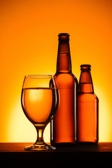 close up view of bottles and glass of beer on surface on orange background