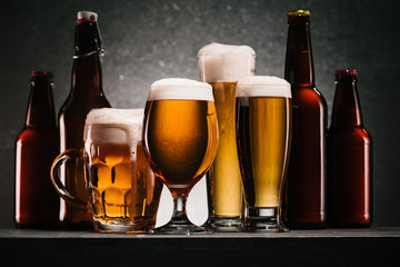 close up view of bottles and mugs of beer with froth on grey background