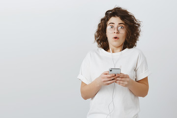 Girl unxpectadly beat record in game. Shocked stunned attractive young female with short curly hair, wearing trendy glasses and casual outfit, holding smartphone, gasping from surprise and anxiety