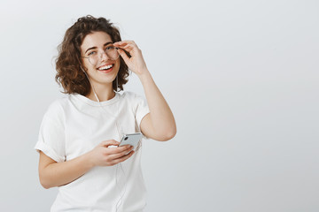 Fototapeta premium You cannot notice girls happiness. Portrait of joyful attractive curly-haired woman, holding rim of glasses and smiling broadly, listening music in earphones and messaging with smartphone