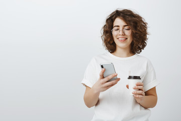Busy cute businesswoman checking schedule. Portrait of satisfied successful designer in glasses, holding cup of coffee and holding smartphone, smiling while looking at screen happily