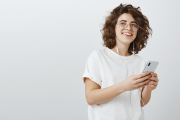 Fototapeta premium Creative female blogger thinking up post to impress followers. Dreamy girl in trendy glasses, looking aside, picturing or recalling nice moment, listening music in earbuds and holding smartphone