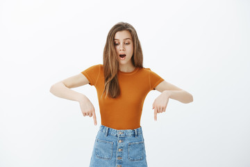 Intense intrigued attractive woman with fair hair, looking and pointing down with dropped jaw, seing something amazing and impressive, saying wow and standing over gray background shocked