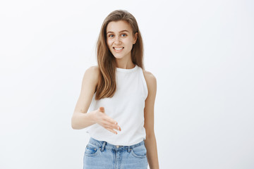 Indoor shot of pleasant good-looking feminine girl with fair hair, pulling palm towards camera to give handshake and smiling cheerfully, greeting business partner and standing confident over gray wall