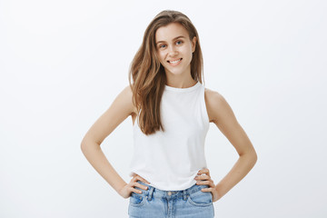 Obraz premium Confident attractive feminine girlfriend with fair hair in stylish tank-top, holding hands on hips and smiling joyfully at camera, satisfied with success, having great time while hanging out in pub