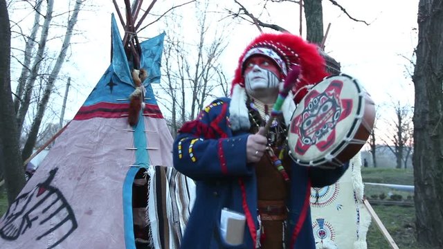 North American Indian in full dress, A man in a suit Indian dancing with a tambourine on the street in front of wigwam