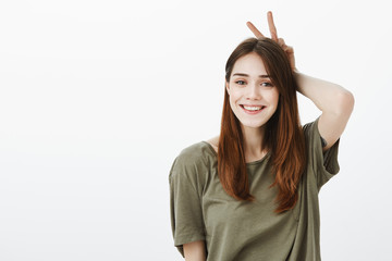 Happy people make positive impression. Portrait of good-looking carefree girlfriend in casual t-shirt, showing v sign behind head, mimicking bunny ears and smiling happilly over gray wall