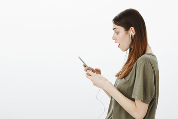 Naklejka premium Woman watching shocking video on smartphone. Attractive stylish girlfriend in t-shirt, holding smartphone and wearing earphones, dropping jaw from interest and surprise while looking at screen