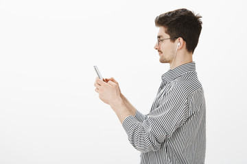 Profile shot of confident attractive bearded guy in trendy glasses and striped shirt, holding smartphone and looking at screen with smirk, messaging with charming girlfriend over gray background