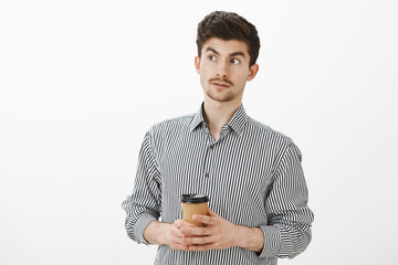 Confused mature european boyfriend with moustache and beard, looking away with lifted eyebrows, drinking coffee and being questioned with behaviour of friend, thinking he strange over gray wall