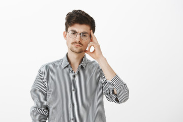 Portrait of serious focused male coworker in round glasses, looking down and holding temple with index finger, concentrating while thinking, making up plan how to avoid uncomfortable situation