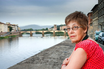 Fototapeta na wymiar Mature tourist woman travelling in Italy stays against bridge over Arno river in Florence.