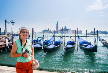 Obraz na płótnie Canvas Mature tourist woman travelling in Italy stays against row of gondolas moored by Saint Mark square against San Giorgio di Maggiore church in Venice.