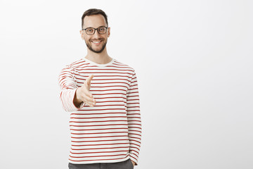 Nice to meet you. Portrait of friendly confident adult businessman in striped pullover, pulling hand towards camera in handshake and smiling joyfully, standing over gray wall, welcoming customer