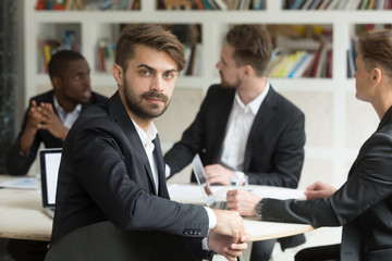 Young talented male worker looking at camera sitting at business meeting. Managers discussing company strategies and plans at background at financial briefing. Diverse team coworking, analyzing ideas