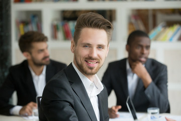 Portrait of young business worker smiling looking at camera at company briefing. Successful team leader posing at corporate meeting with colleagues working behind. Concept of leadership, confidence