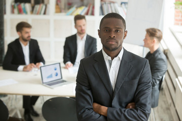 Confident african american businessman looking at camera, work team negotiating behind. Black group leader posing with arms crossed representing business success and achievement