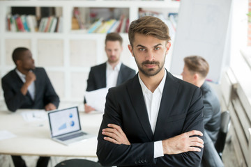 Portrait of caucasian office worker looking at camera standing in foreground, colleagues negotiating in background. Team leader posing leaving subordinates behind. Concept of success and confidence