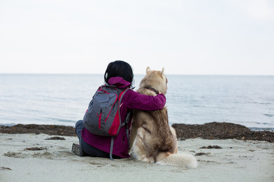 Image of a young girl with backpack and cute siberian husky dog sitting back to the camera and looking at beautiful sea view.