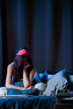 Image of young brunette with insomnia with pink bandage for eyes sitting on bed next to clock