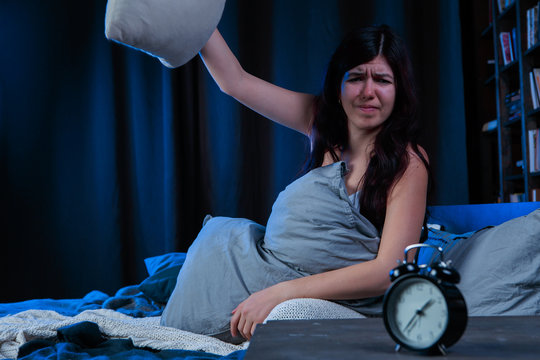 Photo of dissatisfied woman with insomnia throws pillow sitting on bed next to alarm clock