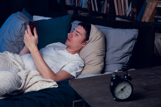 Photo of man with sleeplessness with phone in hands lying in bed next to alarm clock