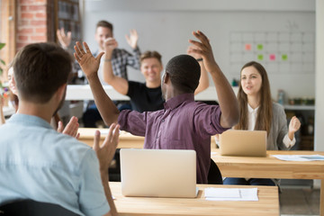 Obraz premium Excited african american raising hands happy about company business success, big achievement and looking back at colleagues reaction. Coworkers celebrating win in modern coworking office