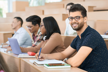 Sideview of Arabian student looking at camera sitting in university during lesson near other students-groupmates. Looking calm, educated and concentrated. Guys reading, studuing, learning, writing.