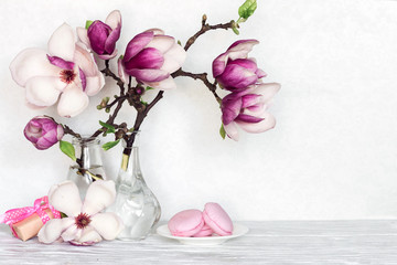 still life with bouquet of pink magnolia flowers in bottles with gift box and macaroons on white table - obrazy, fototapety, plakaty