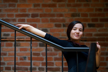 Indoor portrait of young beautiful model of mixed race dressed in black dress standing at the staircase of brick building