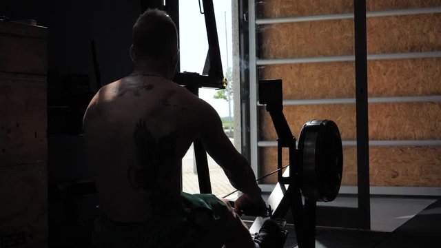 Muscular young shirtless man, training back on equipment, pulling cable at rowing machine. Seen from the back