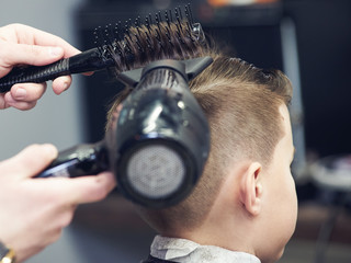 Barber is using hairdryer and hairbrush to make a styling to a Caucasian boy.