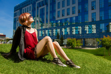 A young hipster girl sits on a green hill in the city.