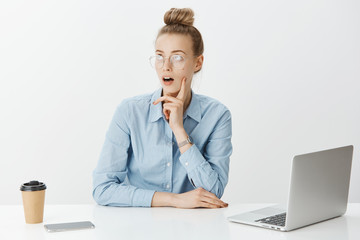 Indoor shot of dreamy creative female coworker with blond hair in glasses, having idea or plan while sitting in office, writing work in laptop, drinking coffee and making notes in smartphone