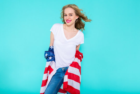I love America. Happy young smiling woman in jeans and white Tshirt holding American flag and looking at camera while standing against pastel blue background.