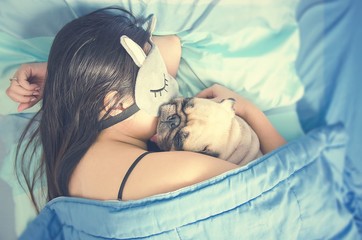 Young woman is lying and sleeping with cute pug dog puppy in bed. Nap with hugging dog
