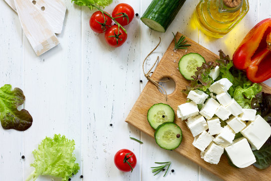 Cooking qreek salad with fresh vegetables, feta cheese and black olives on a white wooden table. Copy space, top view flat lay background.
