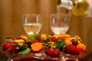A festive chocolate cake with fresh fruits, strawberries, kumquat. In the background, two glasses of white wine are poured from the bottle.