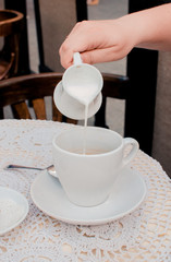 Young caucasian woman pouring milk into morning coffee during breakfast.