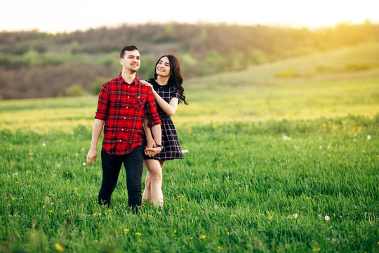 Beautiful couple smoling and holding their hands walking at the green blooming field at sunset. Outdoor