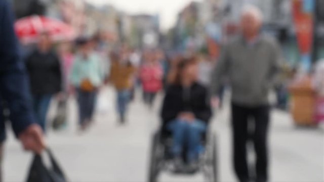 Anonymous disabled people in wheelchair on street. Blurred lady in wheelchair being pushed along crowded city street. Out of focus background from city with people in weekend