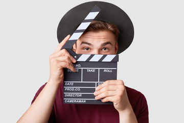 Professional male actor ready for shooting film, holds movie clapper, prepares for new scene, wears special clothes, isolated on white background. Handsome young man poses with clapboard in studio - obrazy, fototapety, plakaty