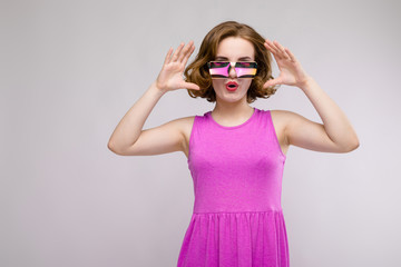Charming young girl in pink dress on gray background. Cheerful girl in square glasses.