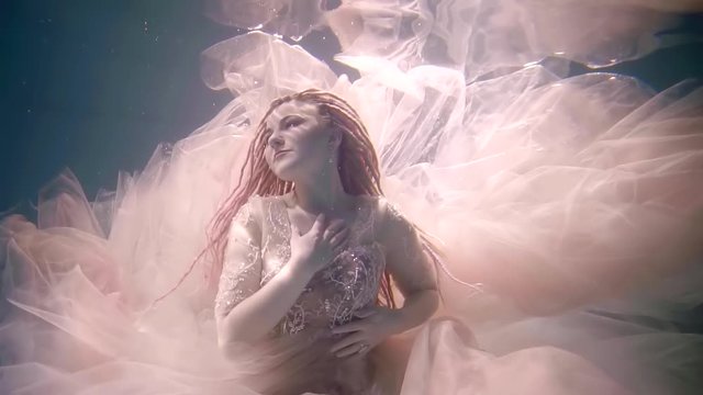 A young woman with dreadlocks on her head and looking like a fairy-tale character is under water