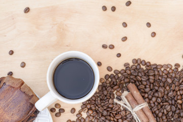 white cup with coffee on a wooden background with coffee grains and cinnamon sticks with a piece of bun