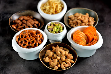 Different kinds of snacks - chips, salted peanuts, cashews, almonds and pistachios, pretzels with salt, potatoes, salted straw.
